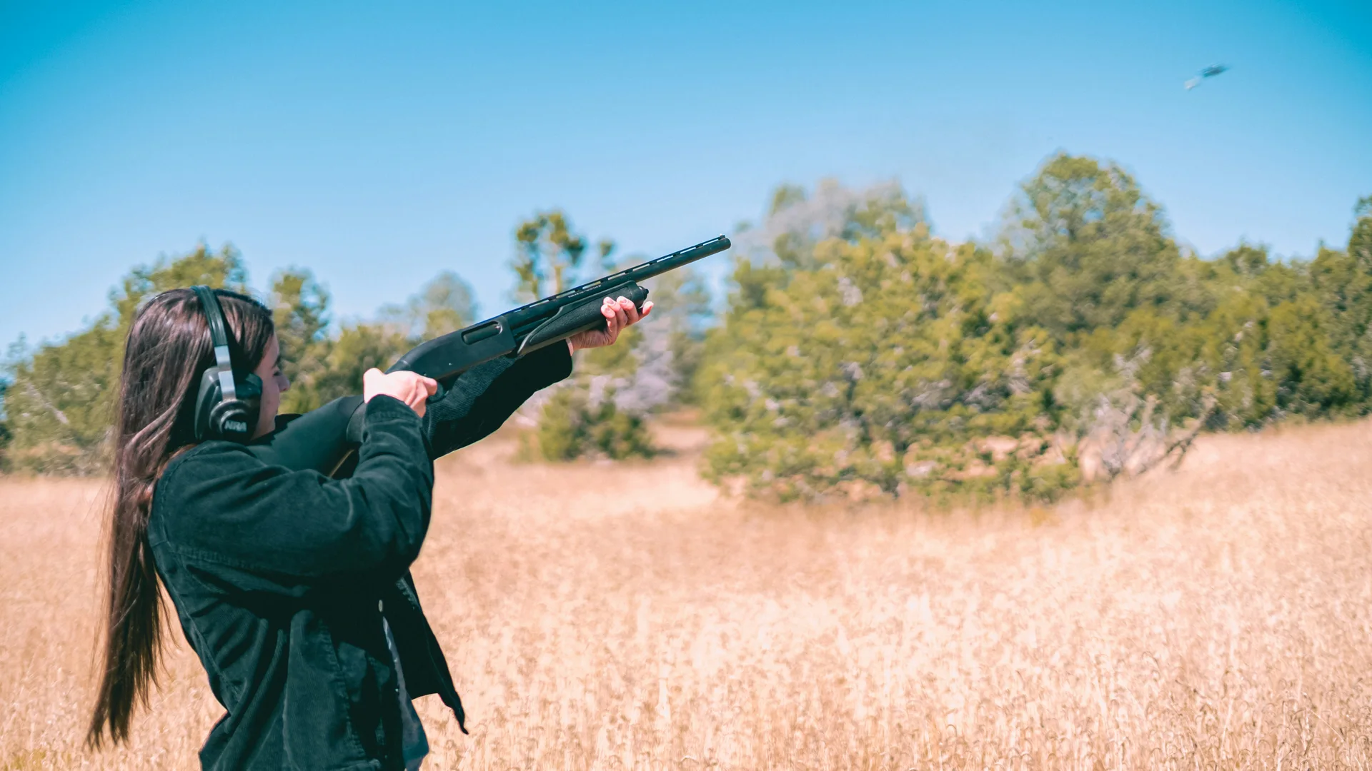 A participant wearing ear defenders aiming a shotgun outdoors